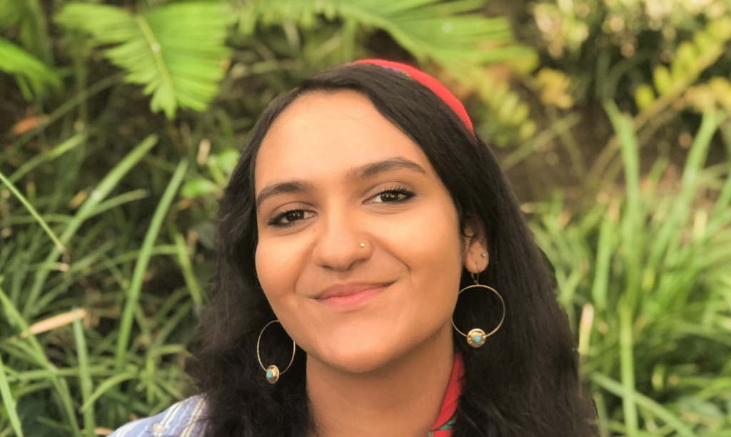 Photograph of Saranya, a 19 year-old student. Saranya has long dark hair, wears gold hoop earrings with little baubles on them, and is smiling at the camera. 