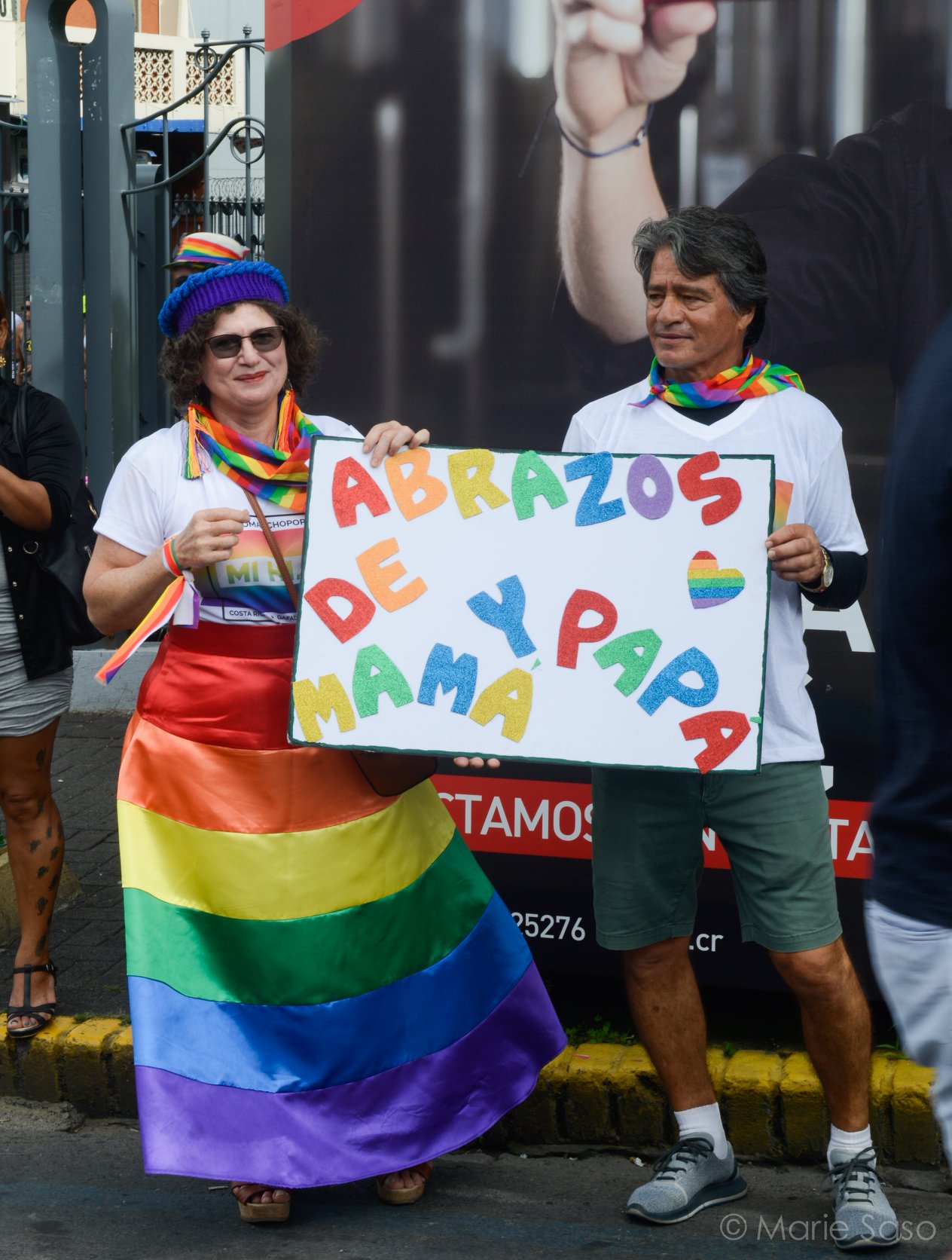 Image shows two people wrapped in pride flags, holding a sign that reads 'Abrazos de y papa mama'