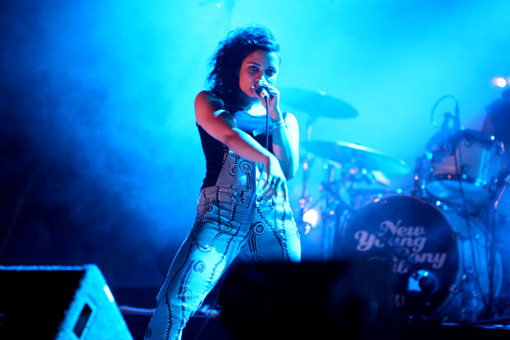 A photograph of Tahita Bulmer of New Young Pony Club in dungarees performing on stage with a microphone held to her face.