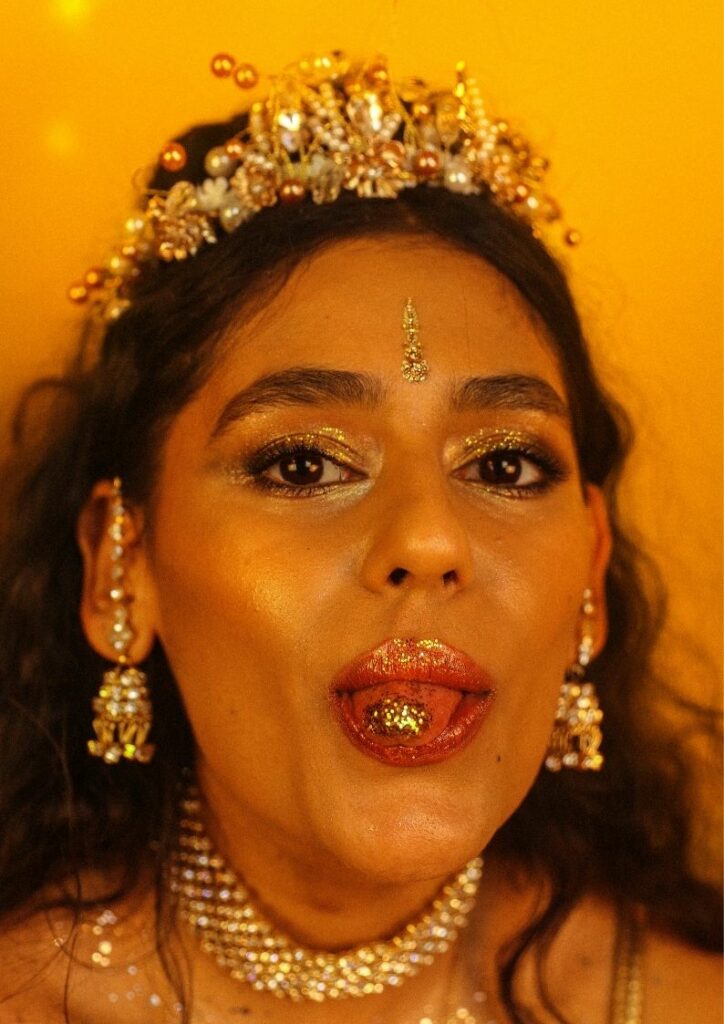 A photo of Muslim punk Nadia Javed from the Tuts. She's wearing lots of gold jewellery and is sticking her tongue out (her tongue has gold glitter on it too). 
