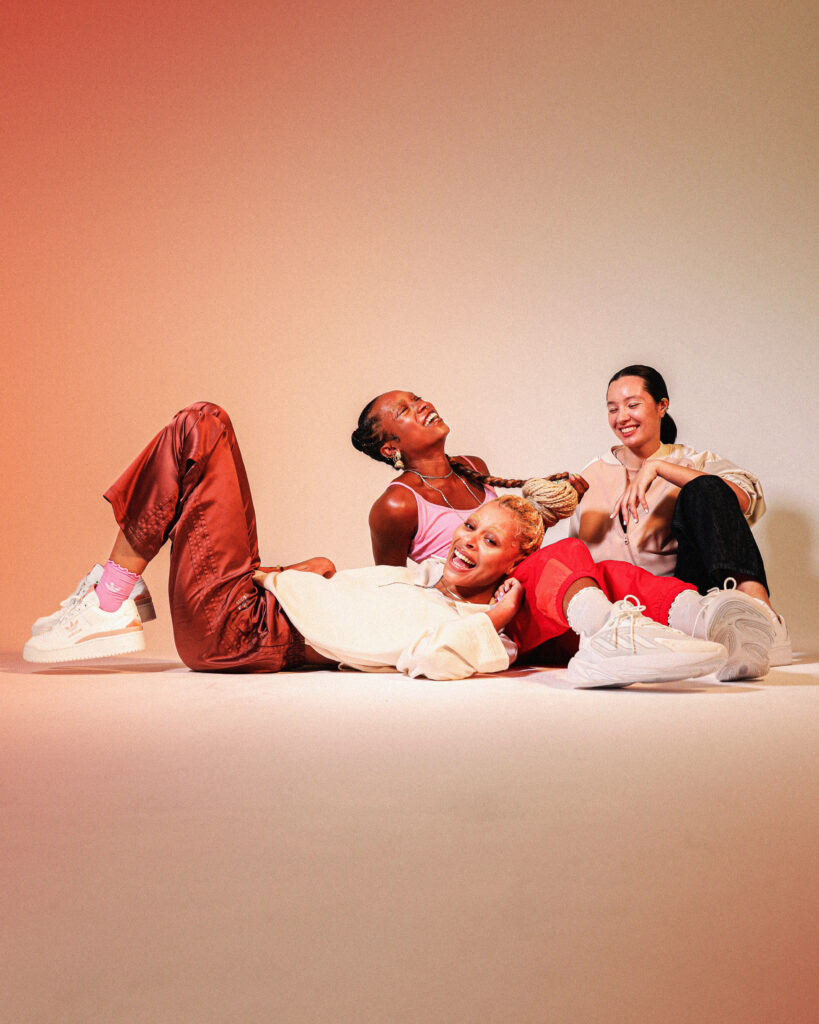 3 models of colour laughing whilst sitting and lying on the floor, in front of a light brown colourama in the adidas 'step into you' collection
