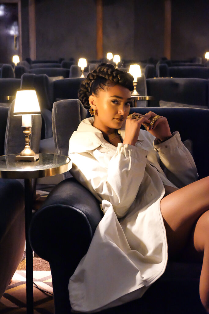 A photo of Joy Crookes seated in a room of plush blue chairs with small glowing lamps. She is wearing a white coat, and her hands are interlinked in front of her face as she looks at the camera.