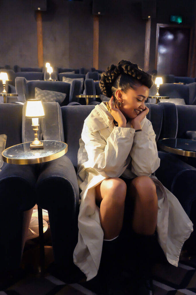 A photo of Joy Crookes seated in a room of plush blue chairs with small glowing lamps. She is wearing a white coat, and her face is cupped in her hands and she is smiling at the ground.