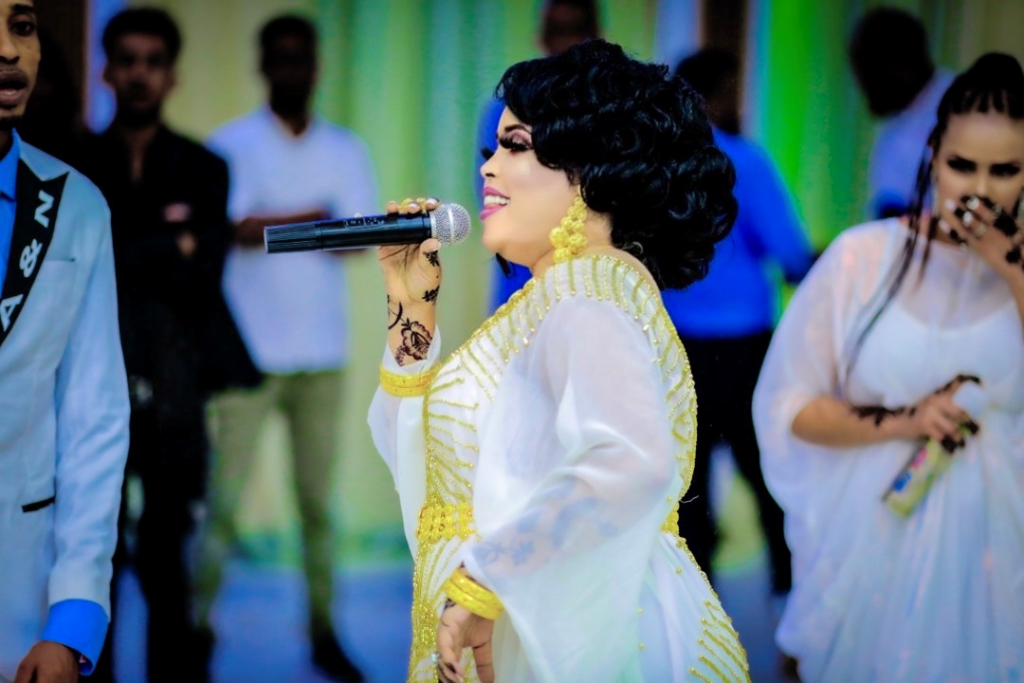A photograph of Nimco Happy in a white and gold outfit, singing into a microphone.