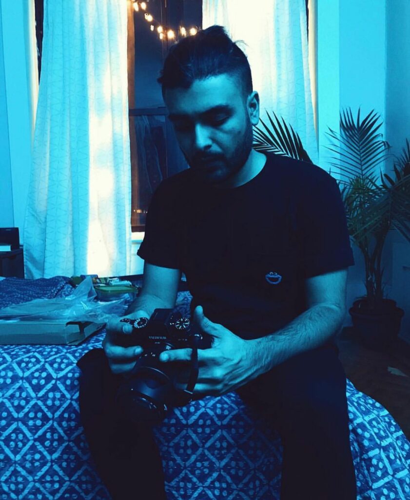 Portrait picture of Akshay sitting on his bed, holding a camera. He is wearing a t-shirt and jeans. There are lights in the background. The picture has a blue filter.