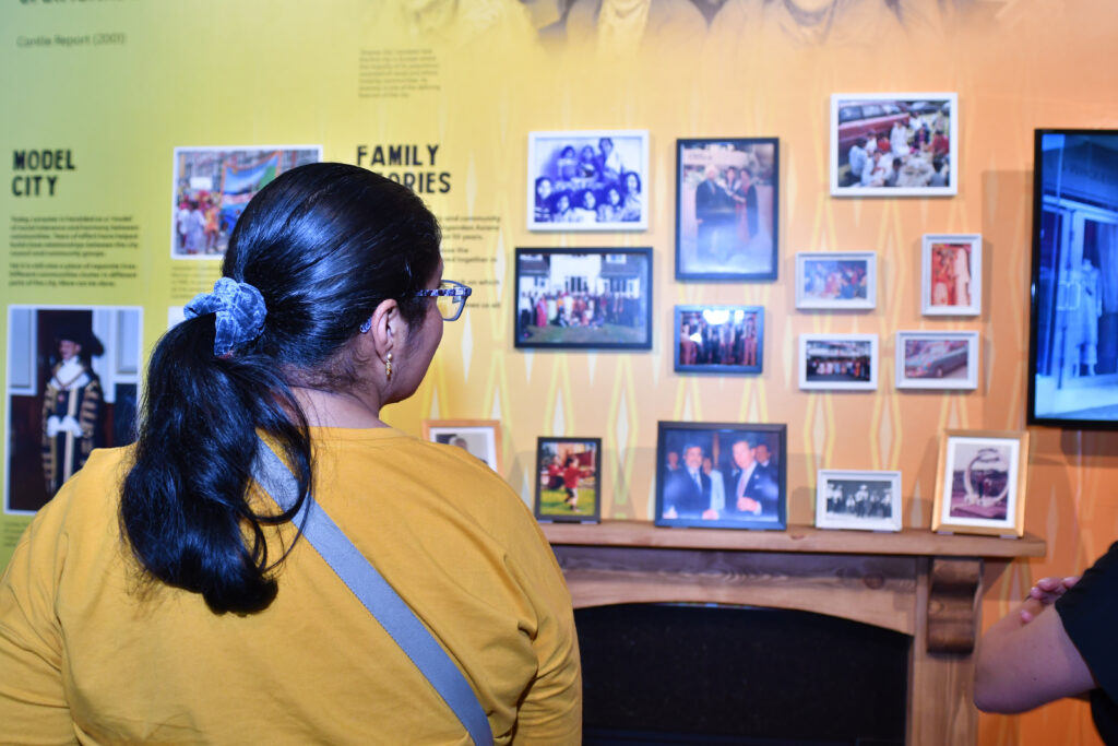 A woman is standing in front of an exhibition. She is looking at photos in frames - the photos are of family memories and Leicester as a city. The photo frames are on an orange background.