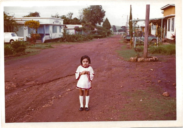 Landscape image of Nisha as a child standing in the middle of a path when in her hometown of Kakira, Uganda. Nisha is wearing a white dress with white socks, with houses in the background.