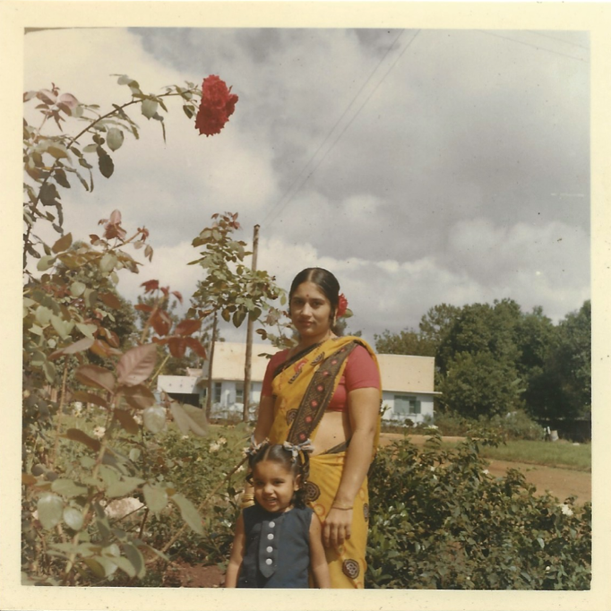 Image of a child (Nisha), standing next to her mother in Uganda. Her mother wears a traditional yellow saree, and they are standing on a field with bushes and trees.