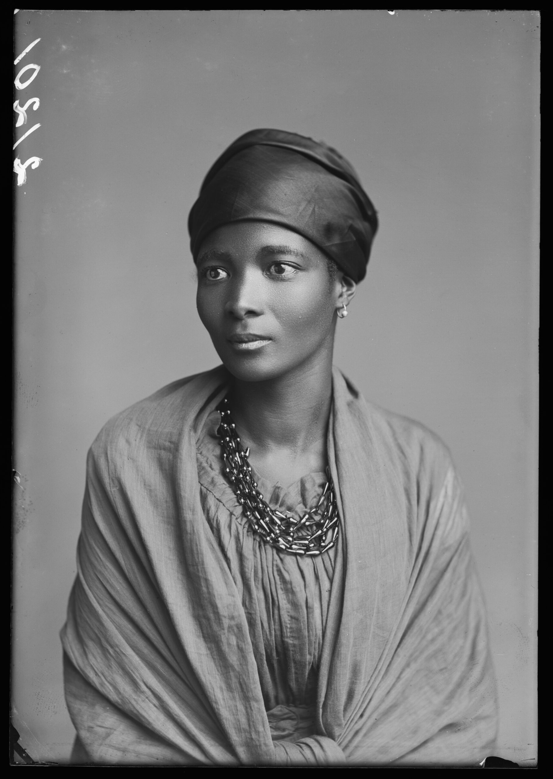 A black and white studio portrait of a woman seated and looking off to the right. She is wearing a headwrap and soft cloths wrap around her
