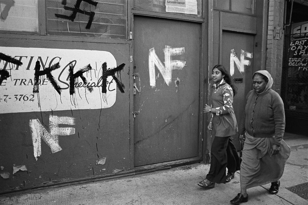 Landscape black-and-white picture of a woman and girl walking past a building with KKK and National Front graffiti.