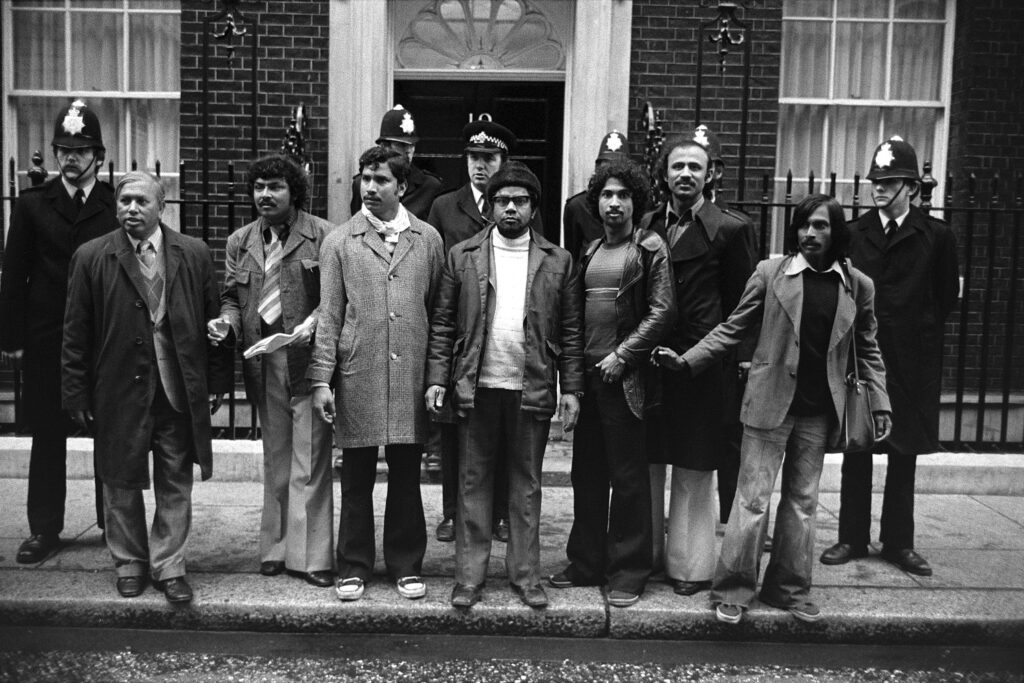 Landscape picture of Bengali men stood outside 10 Downing Street. There are police officers wearing hats standing behind them. The picture is in black-and-white.