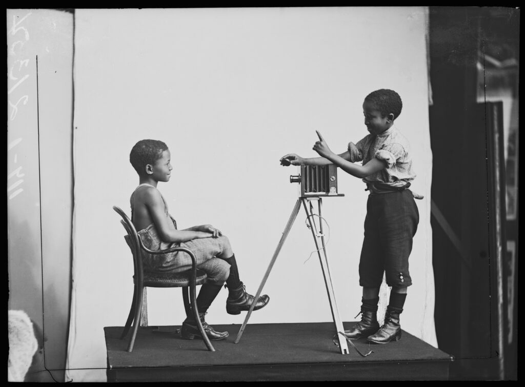 One boy is sitting posing in front of a Victorian camera. The other boy is behind the camera gesturing as if he is about to take a photograph