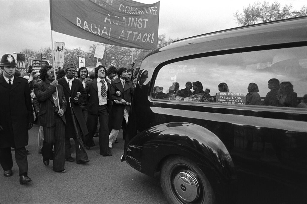 People march behind the car carrying Altab Ali's coffin in support. Some are holding signs supporting the anti-racism movement. There is a police officer on the left, and the picture is in black-and-white.