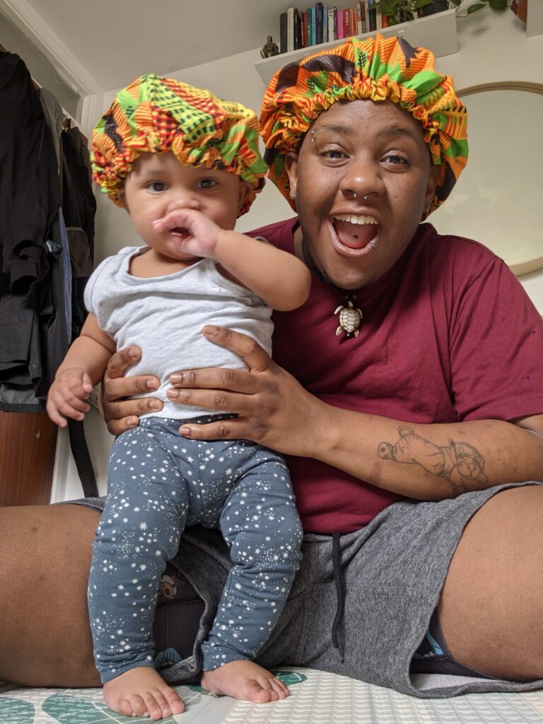 CJ is sitting down holding their child Micah. Both are wearing very cute and colourful bonnets, and looking into the camera