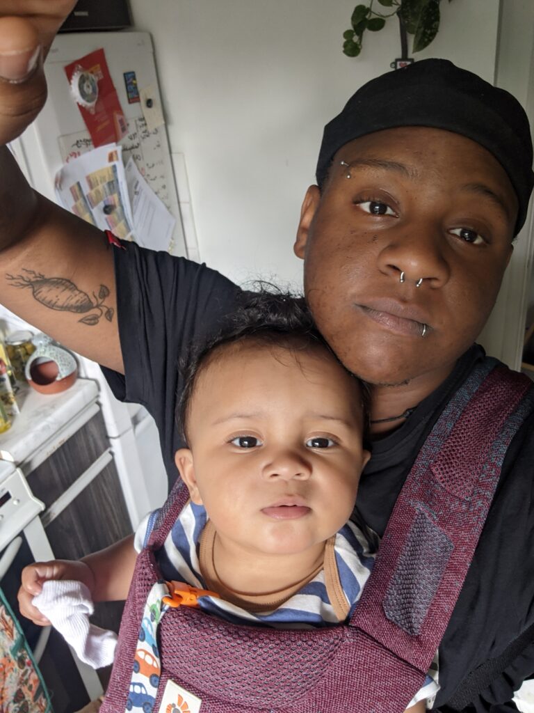 A selfie of CJ and their child Micah in a baby carrier. Both are looking into the camera