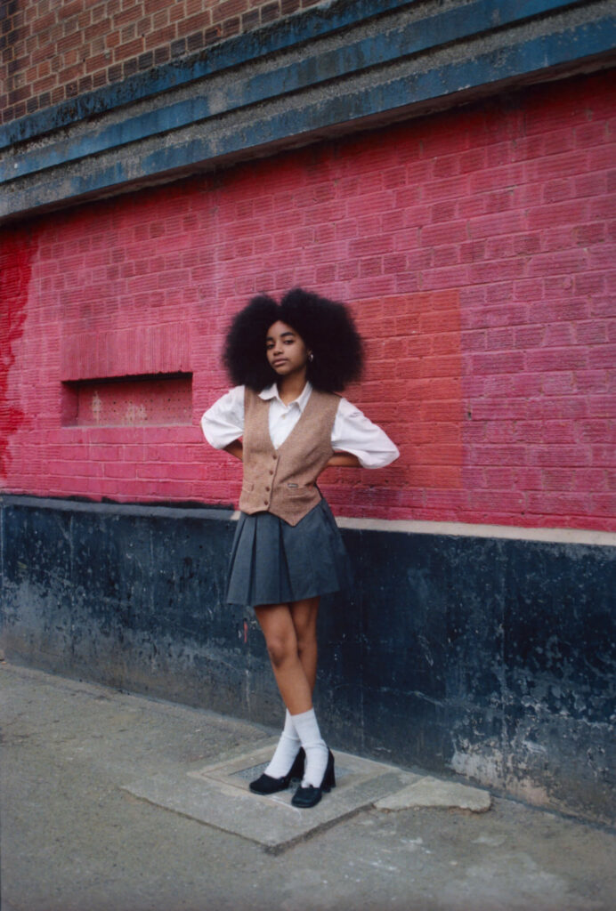 Flowerolove stands with her hands on her waist in front of a red brick wall.