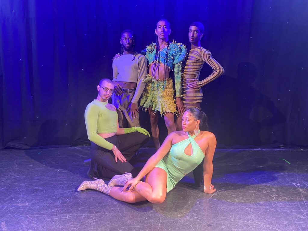 A group of five people are posing, four looking into the camera. Three people are standing up, one is crouching and the other is sitting on the floor in the forefront against the backdrop of a stage and curtain. All are wearing fabulous clothes with neon accents
