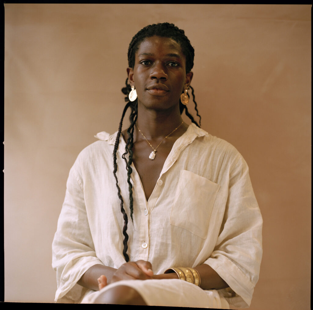 A portrait of Ebun, a black woman who is sitting in front of a beige screen wearing a white shirt, looking into the camera 