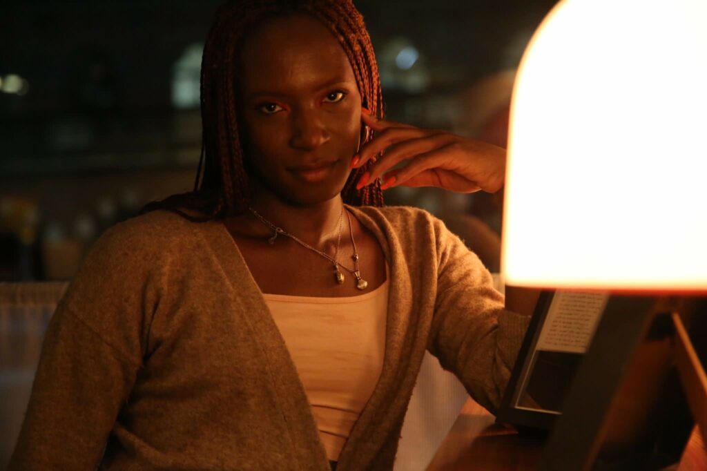A portrait of Chloe, a black woman who is sitting down with her hand to her face, looking into the camera