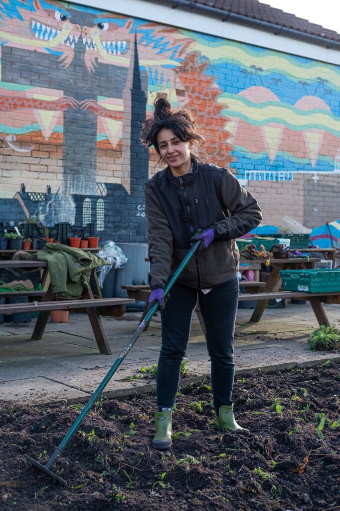 A portrait of Crystal, who is looking into camera and is holding a rake, standing on a patch of soil. Behind Crystal are benches with plant pots and equipment, and a colourful painted mural on a brick wall