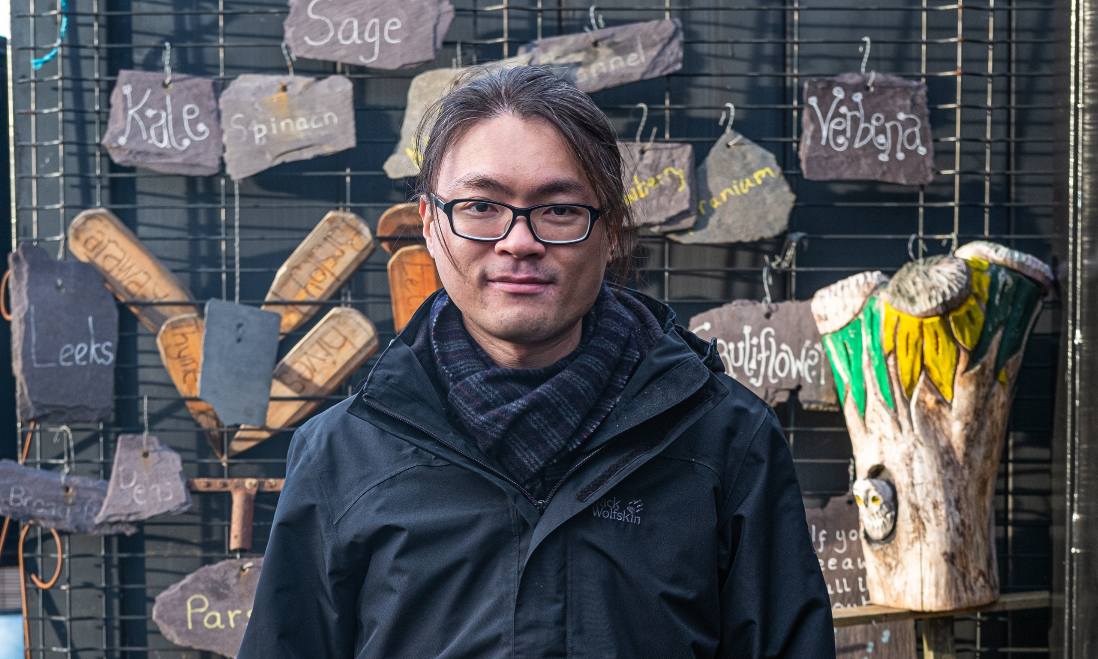 An image of person wearing a coat and looking into the camera. Behind them is a collage of stones with the words of different herbs written on them