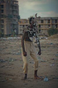 Model stands in Kinshasa surrounding by building work wearing mask made out of disused metal and trash. 