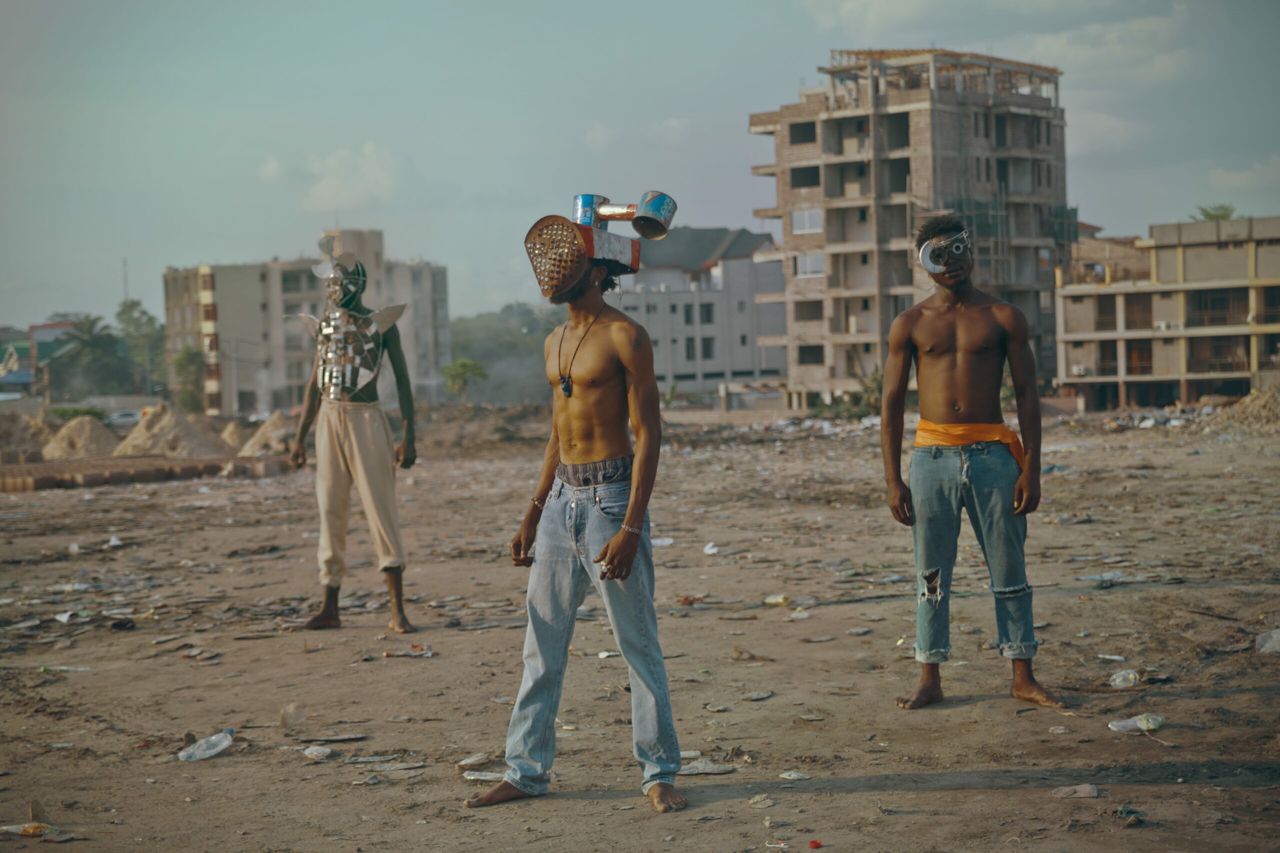 Three models stand in a development site in Kinshasa wearing masks made out of trash.
