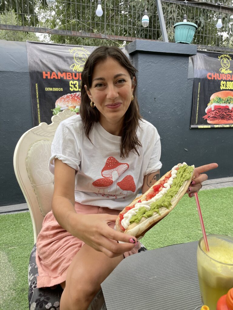 A woman wearing a white t-shirt holds is sitting down, holding a hot dog with red, white and green filling