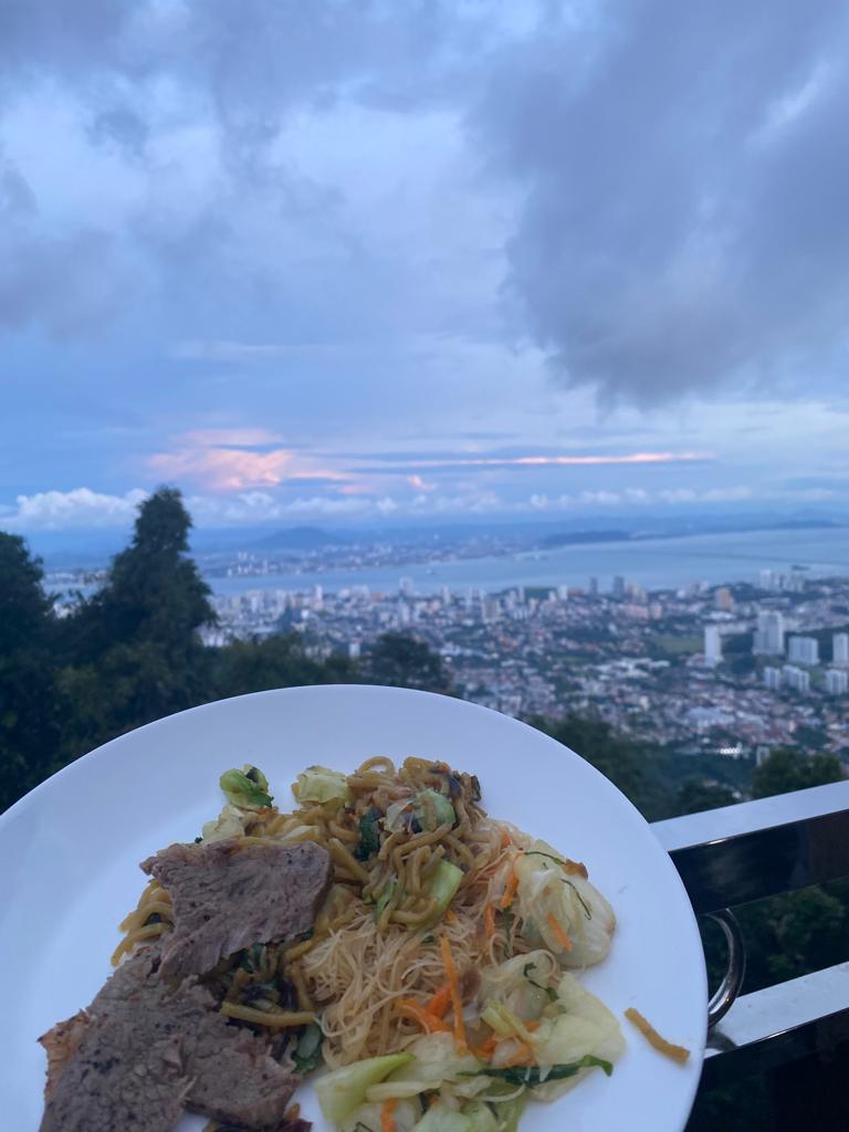 A photograph of a skyline at sunset, with a plat of food including noodles and beef in the foreground