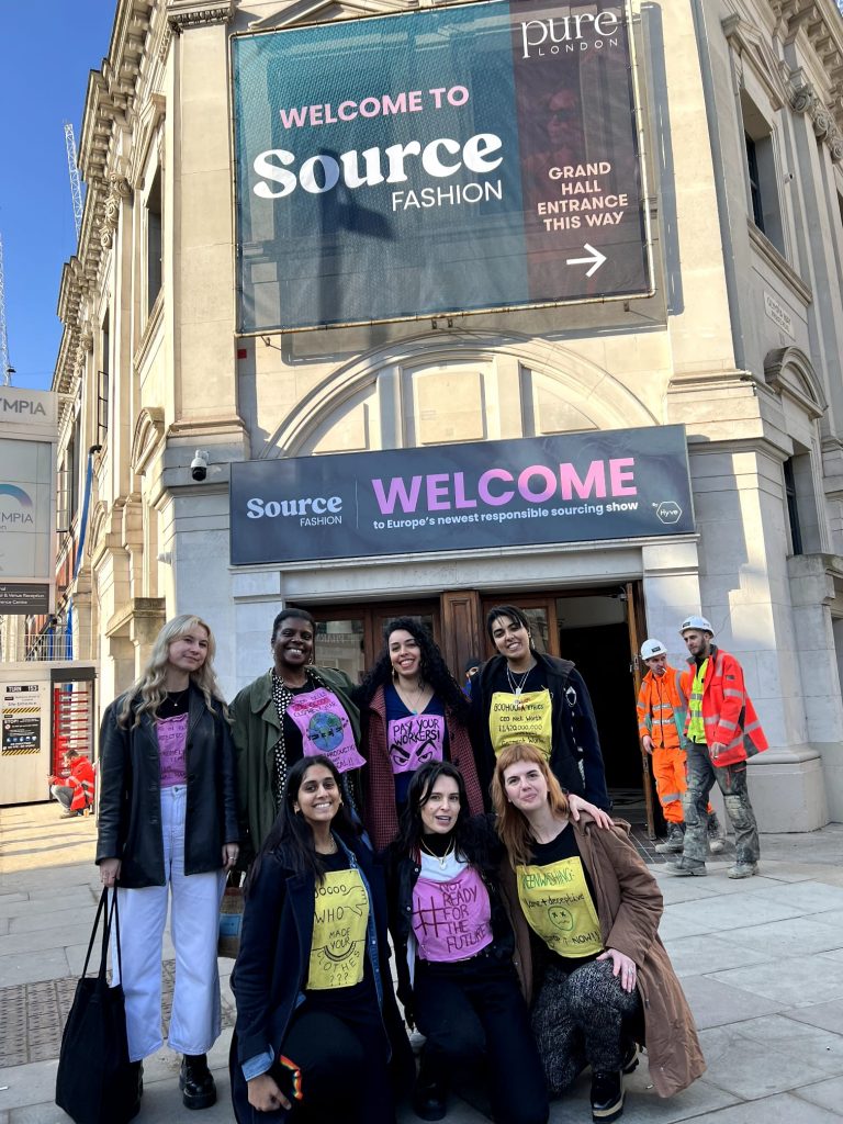 A group of protesters wearing signs posing for a photograph. Behind them is a big advertisement that reads: welcome to source fashion
