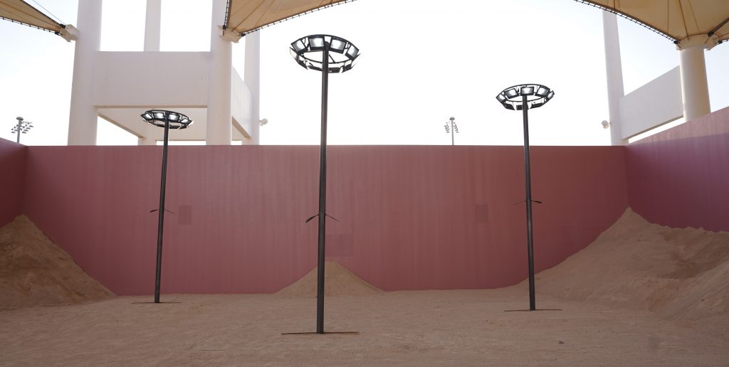 A photograph of an art installation, in an outdoor space, three poles standing in the middle with speakers / lights at the top of them 