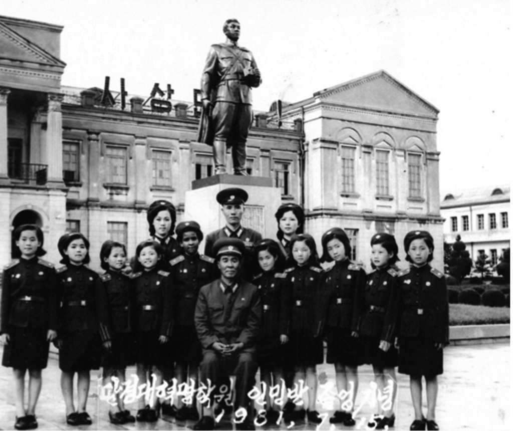 A school class pose for the camera, in front of a statue of Kim Il Sung.