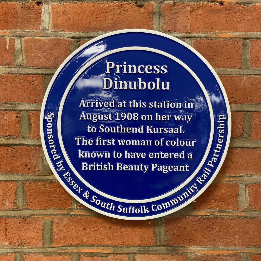 A photograph of a blue plaque with white lettering that reads: Princess Dinubolu, arrived at this station in August 1908 on her way to Southen Kursaal. The first woman of colour known to have entered a British Beauty Pageant. Sponsored by Essex and South Suffolk Community Rail Partnership