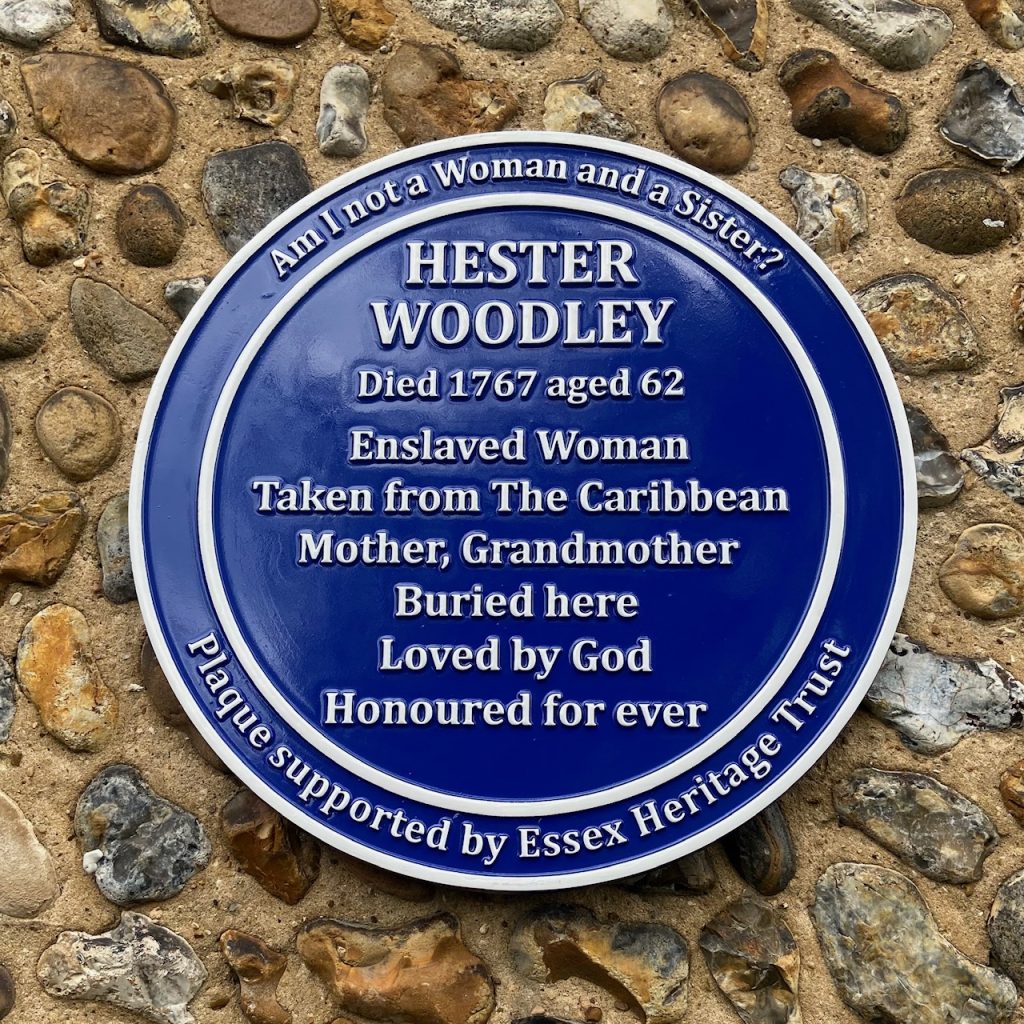 A photograph of a blue plaque with white lettering that reads: Am I not a Woman and a Sister? Hester Woodley, died 1767 aged 62. Enslaved Woman Taken from The Caribbean Mother, Grandmother, Buried here, Loved by God, Honoured for ever. Plaque supported by Essex Heritage Trust